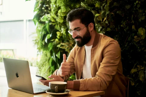 Person is working on an Apple laptop in a cafe, they have a coffee cup beside them and are giving a thumbs up.