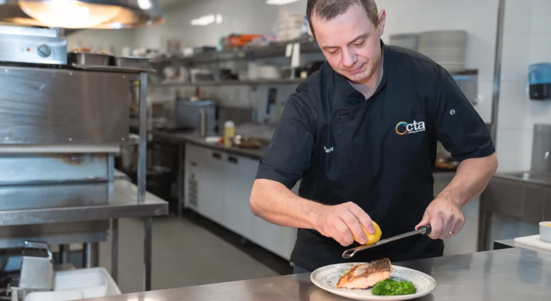 A chef in a commercial kitchen wearing a CTA Training Specialist jacket, he is grating lemon onto salmon
