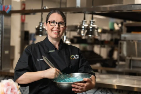 Chef is in a kitchen whisking blue icing in a metal bowl