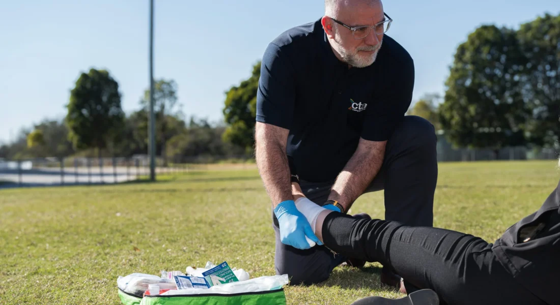 Person is doing first aid on someone's leg while wearing blue plastic gloves. They are kneeling on grass.