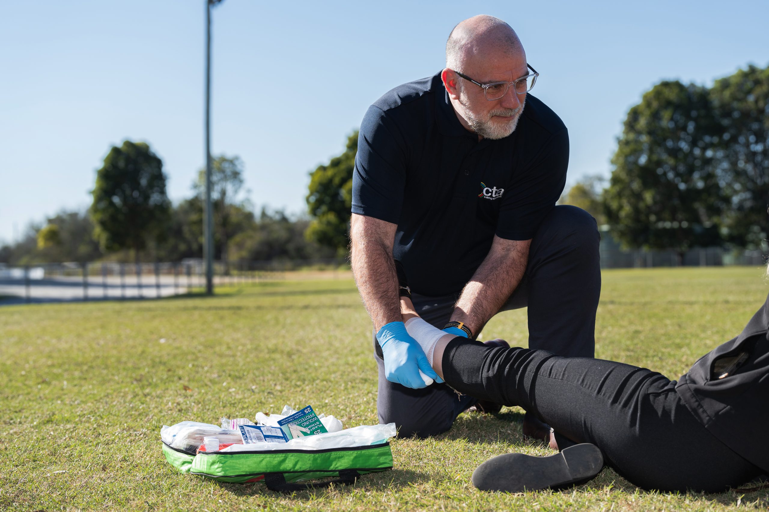 Person is doing first aid on someone's leg while wearing blue plastic gloves. They are kneeling on grass.