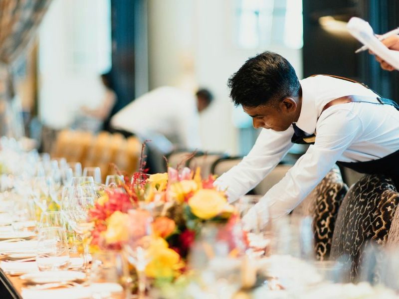 A worker is arranging the table in a nice restaurant. There are yellow flowers on the table.