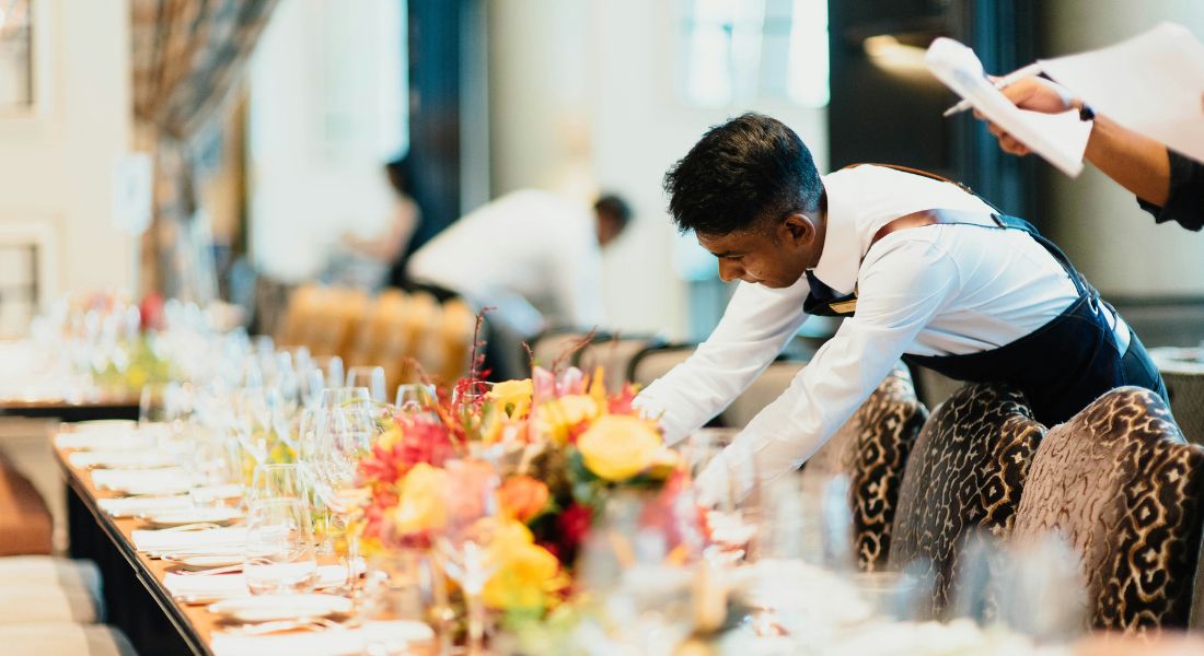 A worker is arranging the table in a nice restaurant. There are yellow flowers on the table.