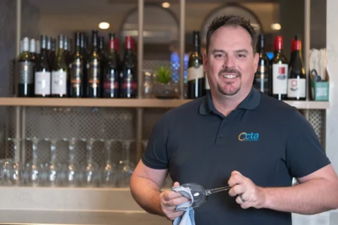 A man standing behind a bar and drying a wine glass, he is wearing a CTA Training Specialist Shirt